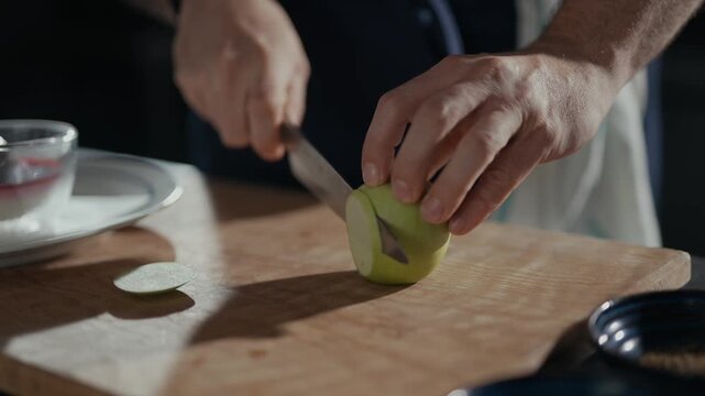 Hands slice green apple with knife on wooden board. Slices fall gently onto cutting surface. Dinner prep scene feels calm and focused. Great for food, cooking, or healthy lifestyle content