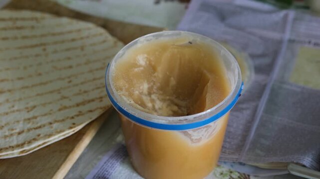 In a simple rural wooden house interior, a plastic bucket of honey stands near flatbread on a table. Small insects are visible around the sweet food, creating a natural but unclean food storage.