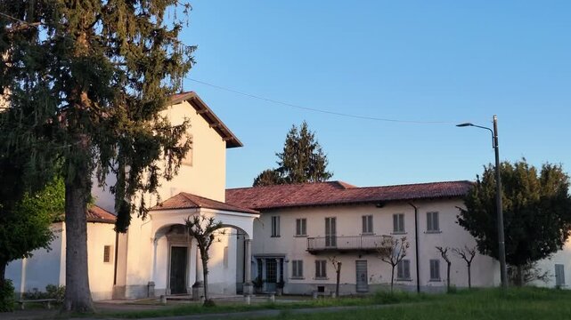 Small Italian church with bell tower in rural village, warm sunlight on traditional architecture surrounded by trees, peaceful countryside scene and cultural heritage landmark.

