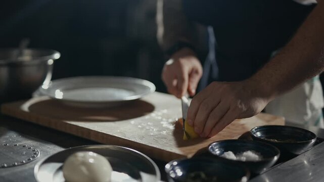 Chef cuts citrus with focused intent. Hands move precisely on a wooden board. Slight steam rises near the cutting surface. Dramatic lighting highlights kitchen tools