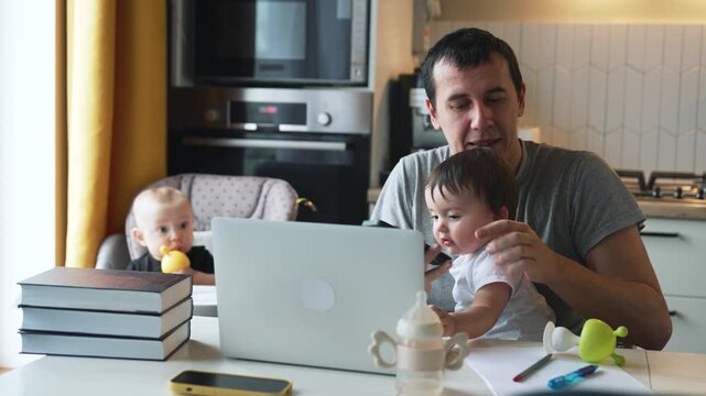 Father multitasking with phone call while holding toddler in kitchen. Dad works on laptop balancing childcare. Parent manages remote work with children. Man juggles parenting and business.