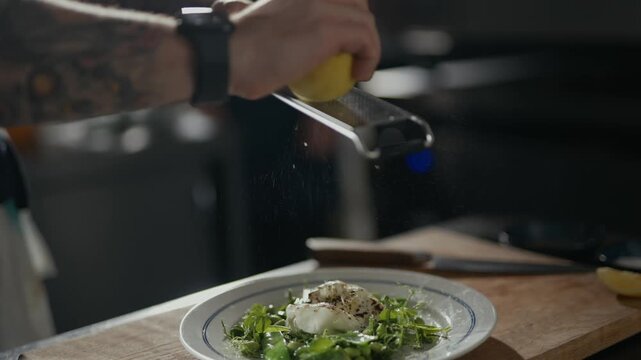 Tattooed hand zests lemon over fresh green salad. Crisp zest falls gently onto leafy greens. Close-up shows zesting action and plate. Food preparation scene, likely in kitchen