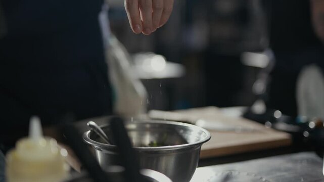 Chef's hand sprinkles salt with care. Focused gesture suggests precision and seasoning. Dark kitchen background highlights culinary action. Ideal for food preparation, restaurant, or cooking scenes