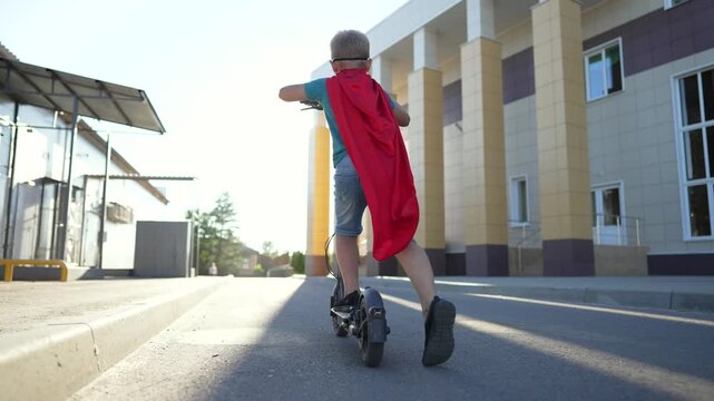 Boy in superhero cape riding electric scooter on sunny street. Child plays hero while riding on urban road. Kid on scooter moves past building in sunlight. Cape flows behind boy on electric ride.