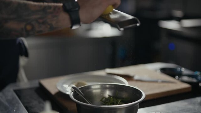 Tattooed chef grates lemon zest over greens. Close-up shows hands and grater in motion. Dramatic lighting highlights culinary action. Ideal for food, restaurant, or cooking content