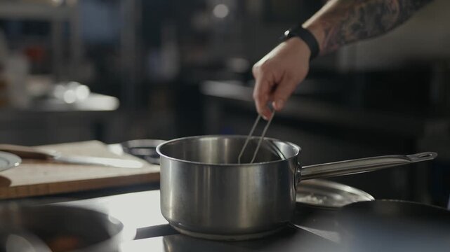 Hand holds strainer with dough over steaming pot. Cook's tattooed arm guides dough carefully. Steam rises as pot heats on countertop. Scene suggests skilled culinary preparation