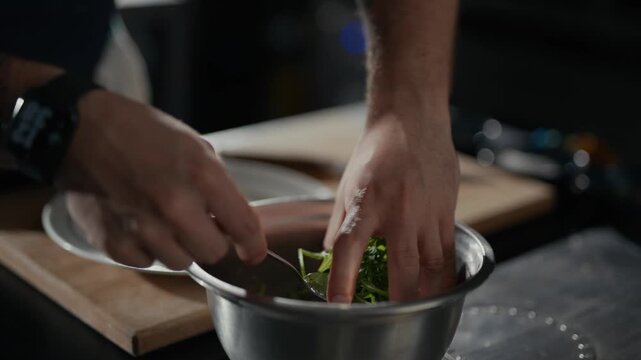 Hands mix greens in a stainless steel bowl. Dark kitchen setting with wooden cutting board. Calm, focused cooking action with no visible emotion. Ideal for food prep, health