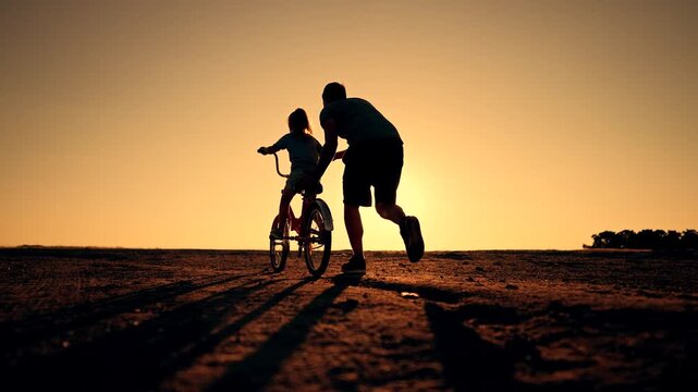 Father teaching daughter to ride bicycle at sunset. Dad helps child learn cycling on dirt field. Parent runs beside girl on bike. Family bonding during golden hour. Father and daughter silhouette.