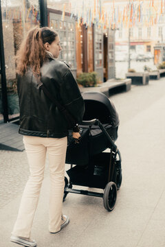 A woman in a leather jacket walking along the sidewalk and pushing a black stroller in the urban space.