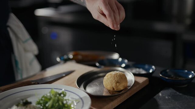 Hands gently shape dough on a wooden board. Plates hold greens and grains beside metal bowls. Dark kitchen hints at quiet culinary creation. Perfect for food prep, cooking tutorials