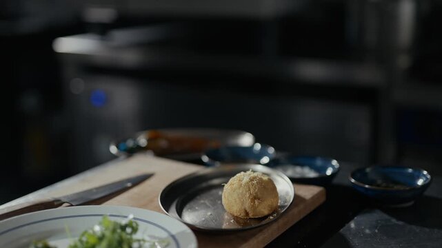 Tattooed hand grates fresh garlic over dough. Dough rests on plate beside greens and tools. Kitchen backdrop suggests focused, hands-on preparation. Ideal for culinary tutorials or food prep sequences