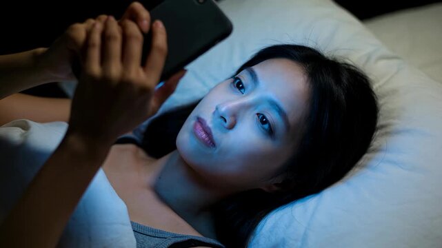 Asian woman using smartphone lying on bed at night. Girl scrolling mobile screen in dark bedroom. Digital connection and insomnia concept, night lifestyle scene.