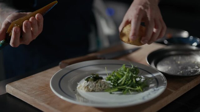 Tattooed chef's hands add vibrant orange garnish. Plated dish features greens, egg, and savory centerpiece. Warm, focused lighting enhances kitchen setting. Perfect for food, culinary