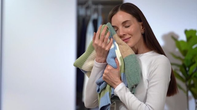 Smiling woman pressing soft sweaters to cheek and stroking knitwear by hand on light wardrobe at home background. Concept of fresh laundry, aroma fabric softener, clothing care and tactile comfort
