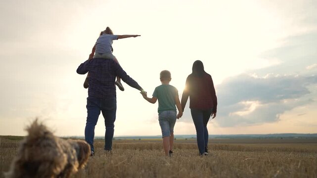 Family walking together in field at sunset. Parent carries child on shoulders while holding hands. Parents and children walk with dog. Family enjoys outdoor walk. Sunset family time in countryside.