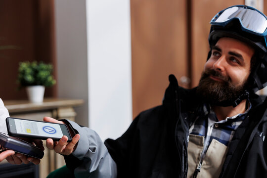 In ski resort lounge area, caucasian man pays with cell phone while staff assists with pos machine. Close-up view of male traveler wearing wintersport outfit using smart device for nfc payment.