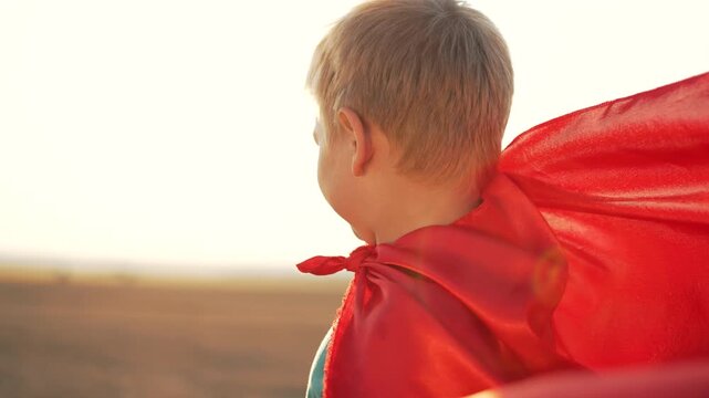 Young boy wearing red superhero cape playing outdoors at sunset. Child pretending to be hero in field. Kid with cape enjoying outdoor play. Boy imagining superhero adventures at golden hour.
