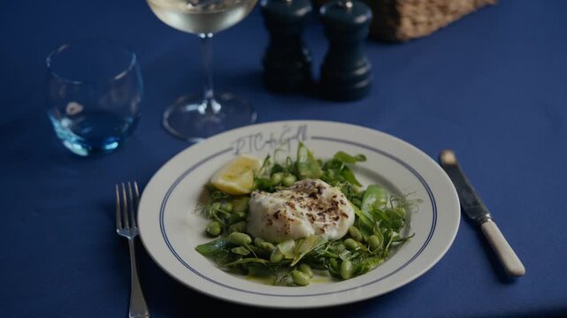 Plate holds fresh greens and a fish fillet. Lemon wedge rests beside the dish. White wine glass and blue water glass sit nearby. Silverware and dark salt and pepper shakers complete the table