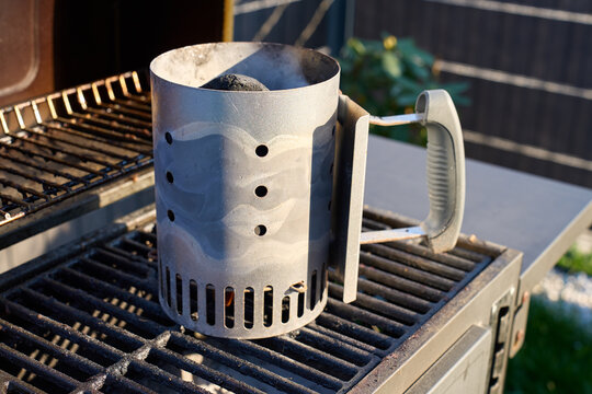 Metal charcoal chimney starter stands on barbecue grill with coals and light smoke