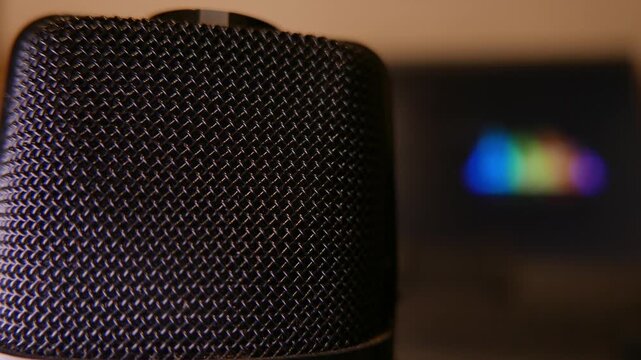 Slow zoom in revealing details of a professional microphone in a recording studio. Blurred colorful sound levels moving intensely on a laptop screen in the background. Close-up of a podcast microphone