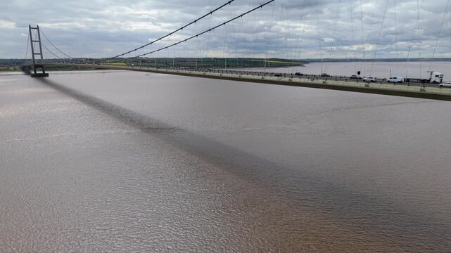 Aerial view of Humber Bridge with vehicles crossing the suspension span over the murky estuary waters under a cloudy sky in Hessle, East Riding of Yorkshire, United Kingdom.