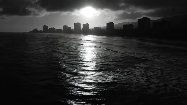 Dramatic black and white ocean shoreline with sun reflection and city skyline silhouetted against dark clouds