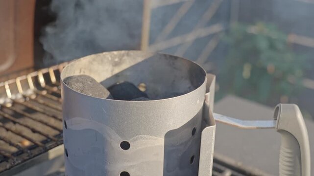 Metal charcoal chimney starter with briquettes and smoke on outdoor grill. Concept of barbecue preparation and outdoor cooking