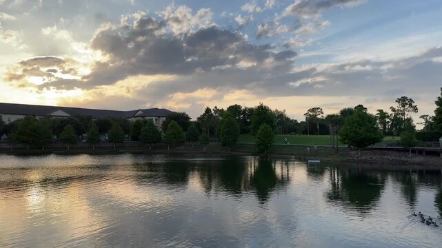 Calm water reflects trees, clouds, and a walking bridge during a quiet sunset moment in a park landscape. The tranquil natural scenery conveys relaxation, travel, and outdoor leisure.