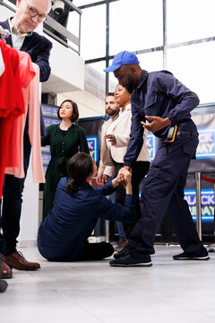 Black Friday shopper injuries. African American guy security guard wearing uniform keeping clothing store and customers safe during busy sales season, helping shopper get up from floor