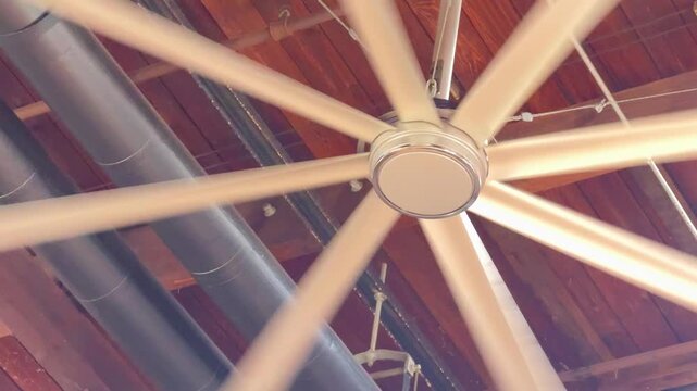 A large ceiling fan rotates rapidly beneath a wooden roof structure. Motion blur and warm indoor lighting create a dynamic mechanical detail scene.