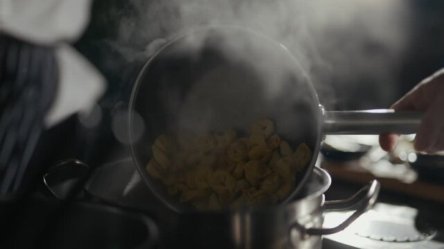 Steam rises from a simmering pot. Cooked pasta pours into boiling water. Hands move with purpose and focus. Heat and steam fill the kitchen. Food preparation in a professional setting