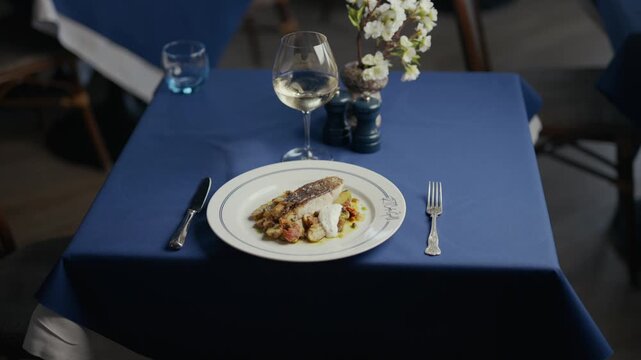 Elegant table set for a fine dining experience. Blue cloth, white plate, wine glass, fork, knife. Floral arrangement adds delicate touch. Still scene conveys quiet anticipation