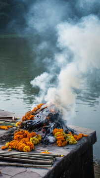 Traditional Hindu cremation pyre with rising smoke on riverside platform, solemn funeral ritual scene with marigold flowers, sacred farewell, remembrance, grief and spiritual memor
