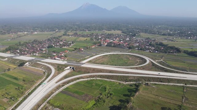 Aerial view of a modern highway interchange surrounded by lush green rice fields and a rural village with Mount Merapi in the distance Klaten, Central Java, Indonesia.