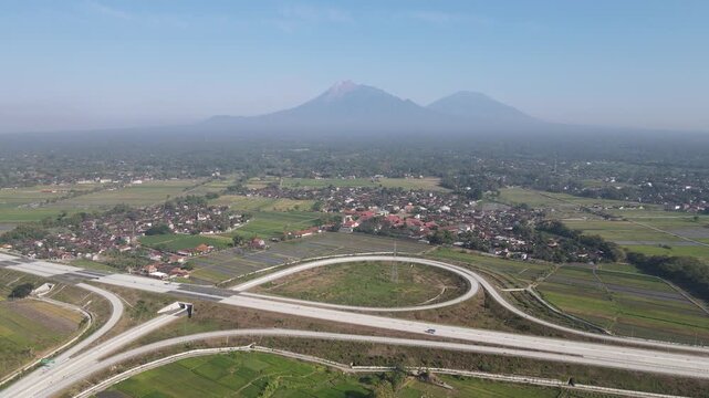Aerial view of a modern highway interchange surrounded by green rice fields and a local village with Mount Merapi in the background in Klaten, Central Java, Indonesia.