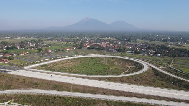 Aerial view of a circular highway interchange loop surrounded by green fields and village houses with Mount Merapi in the background in Klaten, Central Java, Indonesia.