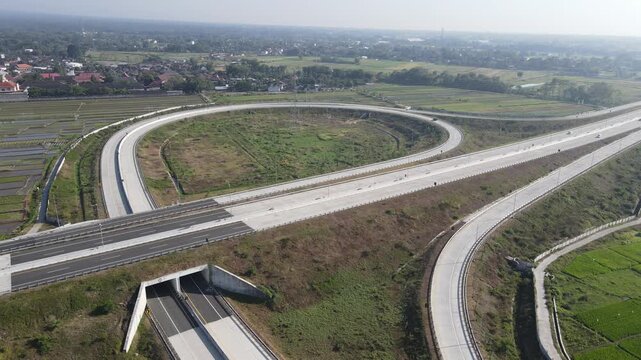 Aerial view of a modern highway interchange with circular ramps surrounded by green rice fields and rural settlements in Klaten, Central Java, Indonesia.