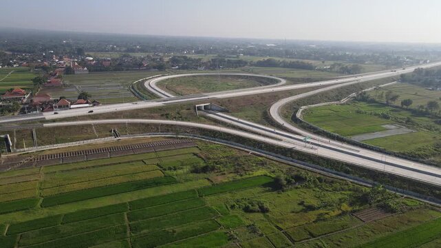 Aerial view of a modern highway interchange with a circular ramp surrounded by lush green rice fields and rural settlements in Klaten, Central Java, Indonesia.