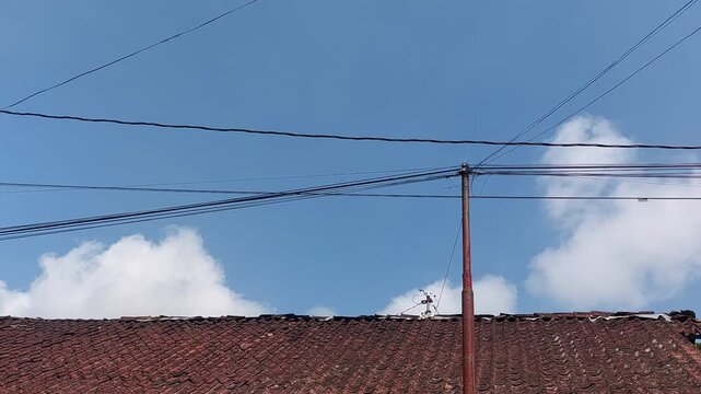 Rustic terracotta tiled roof with an old utility pole and tangled power lines against a bright blue sky, featuring white fluffy clouds, an outdoor scene