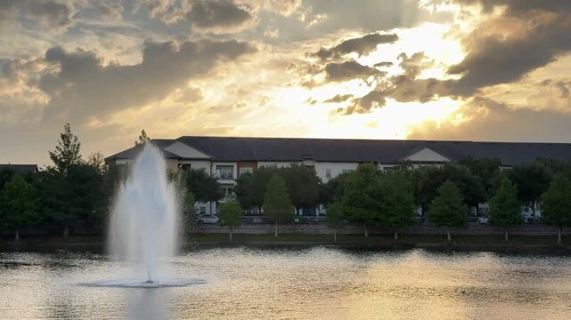 A tall water fountain rises from a calm lake with warm sunset clouds glowing behind nearby buildings. Reflections on the water create a peaceful evening landscape.