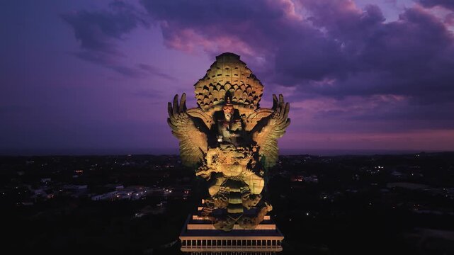 Aerial view of the massive illuminated Garuda Wisnu Kencana statue under a purple twilight sky with city lights in the background in Kecamatan Kuta Selatan, Bali, Indonesia.