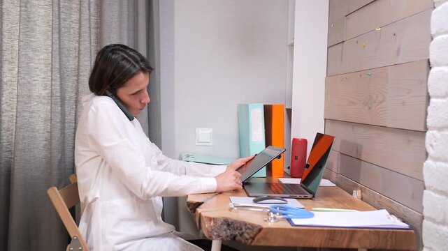 Medical professional in a white lab coat engaging in a virtual consultation, actively communicating on the phone while simultaneously operating a digital tablet and laptop at a rustic wooden desk