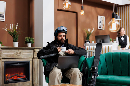 Young man in wintersports gear sits in lounge area browsing on minicomputer and enjoying hot drink. Male traveler holding his beverage while engaged in snowy slopes online research with his laptop.