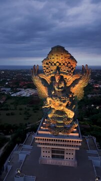Aerial view of the illuminated Garuda Wisnu Kencana statue at dusk under a cloudy sky in Kecamatan Kuta Selatan, Bali, Indonesia.