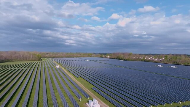 Aerial view of a large solar farm with rows of photovoltaic panels surrounded by green fields and trees under a cloudy sky in Indianapolis, Indiana, United States.