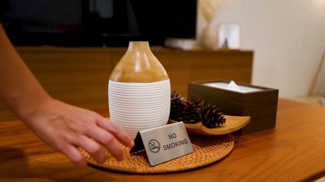 Womans hand placing silver no smoking sign on wooden table next to essential oil diffuser in living room.