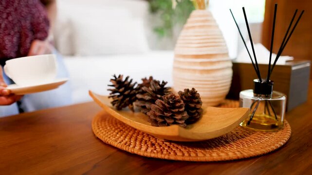 Woman placing white cup of tea on wooden table with decorative pine cones and home fragrance diffuser.
