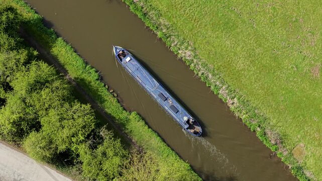 Aerial view of a blue narrowboat cruising along a calm canal surrounded by lush green fields and trees Barton-under-Needwood, England, United Kingdom.