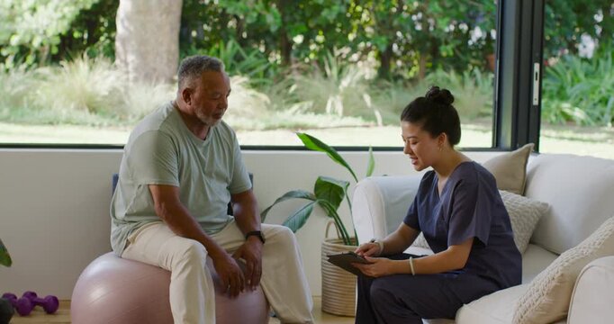 Diverse caregiver in scrubs opening tablet, tapping; senior man shifting on ball at home for rehab