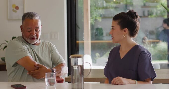 Senior African-American man rubbing arm talking Asian woman calming in scrubs with cell in kitchen